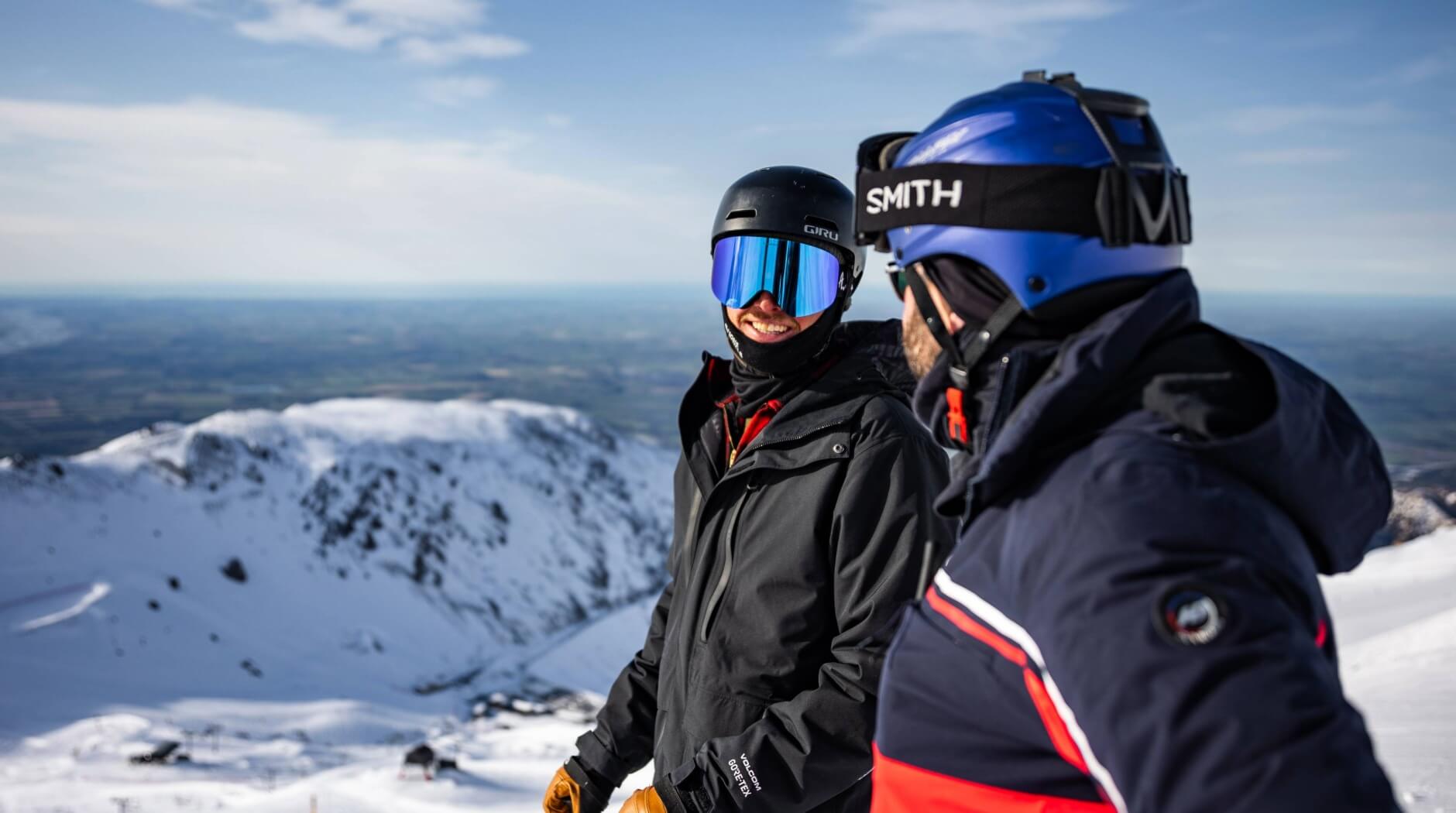 Two skiers in helmets and goggles standing and chatting on a snowy mountain with a clear sky.
