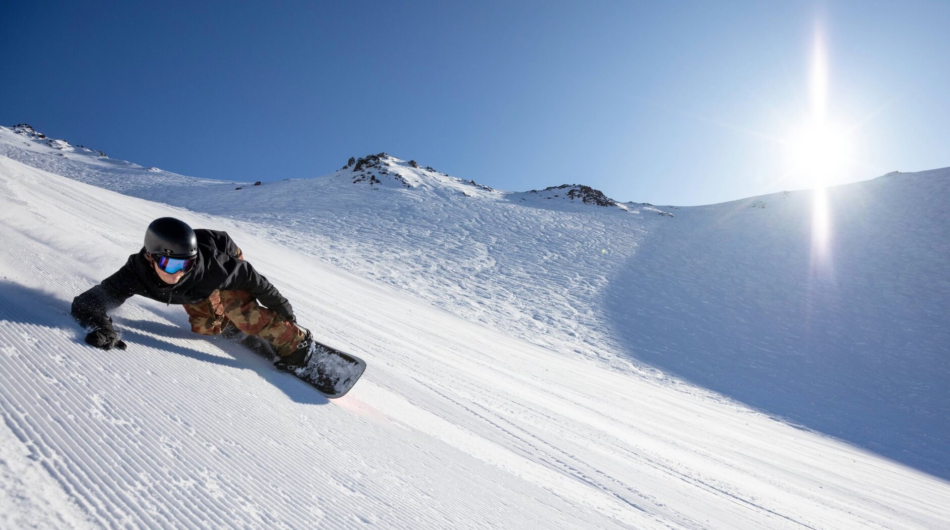 Snowboarder carving sharply on freshly groomed snow on a sunny mountain slope with clear blue sky.