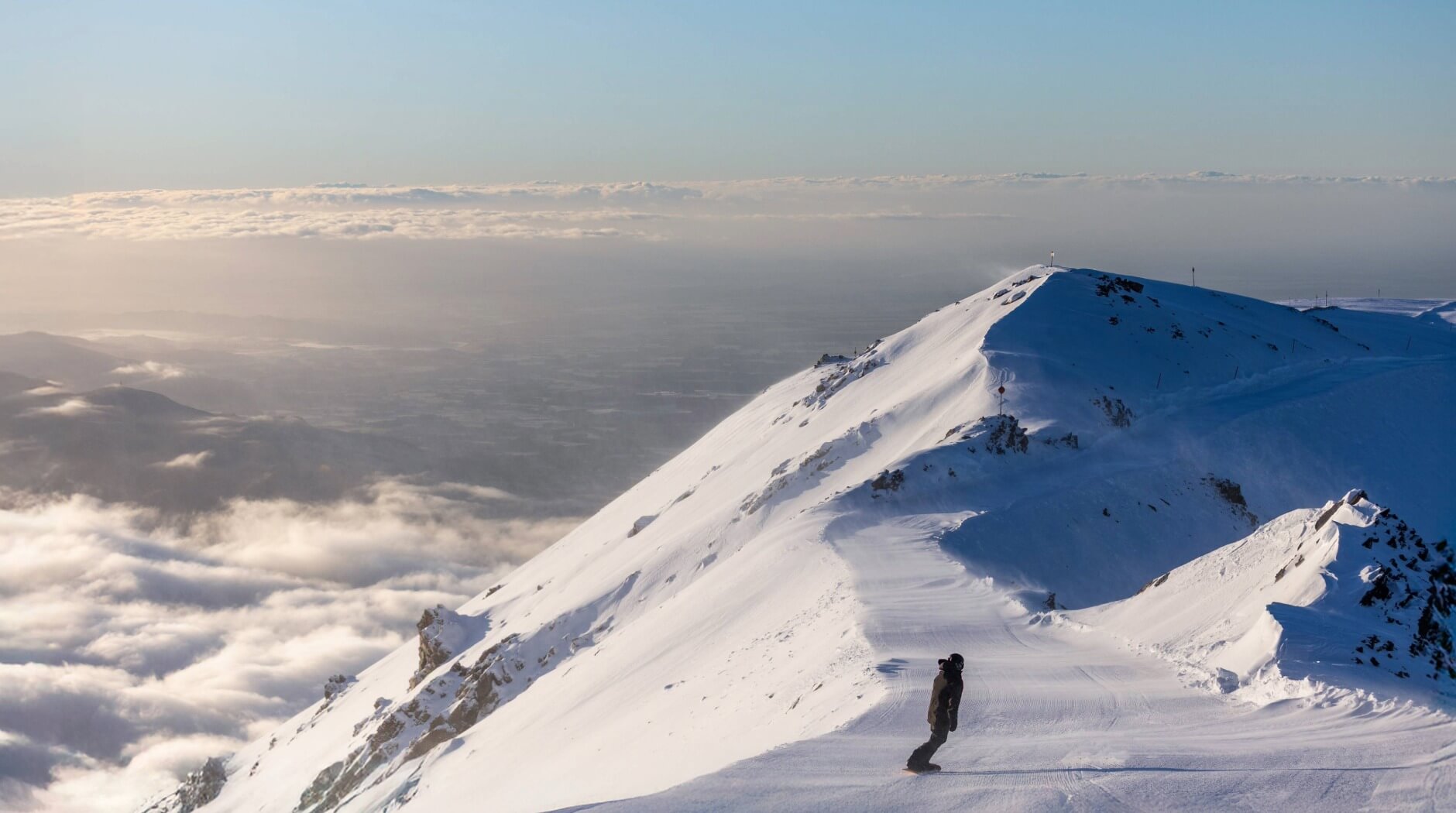 Person snowboarding down a snowy mountain slope with a scenic view of clouds and distant hills.