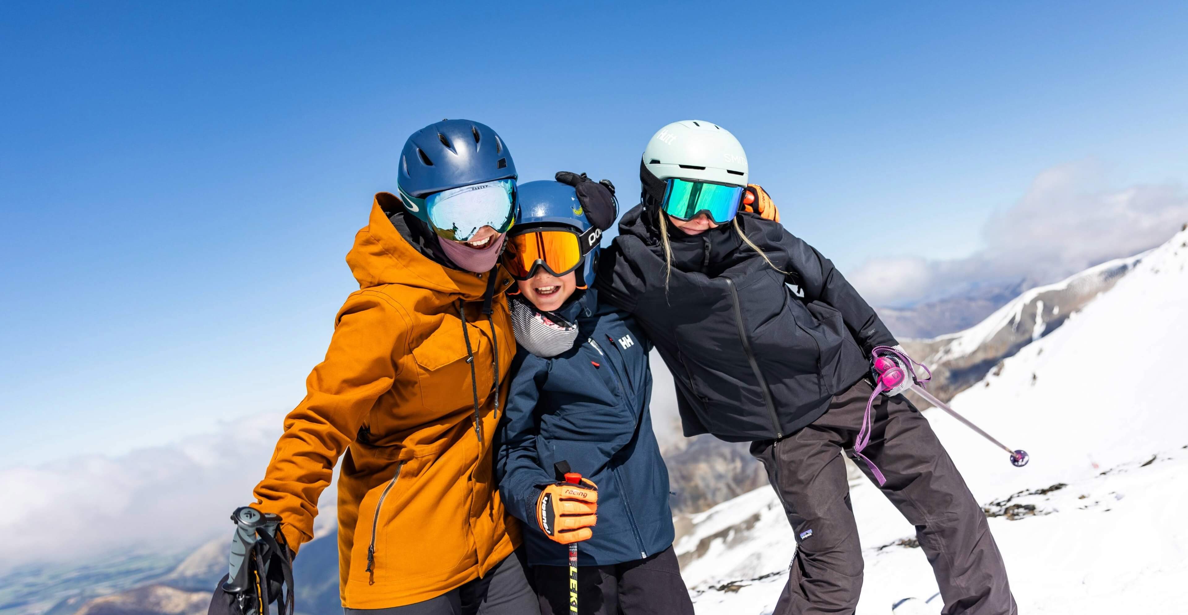 Three skiers wearing helmets and goggles posing together on a snowy mountain under a clear blue sky.