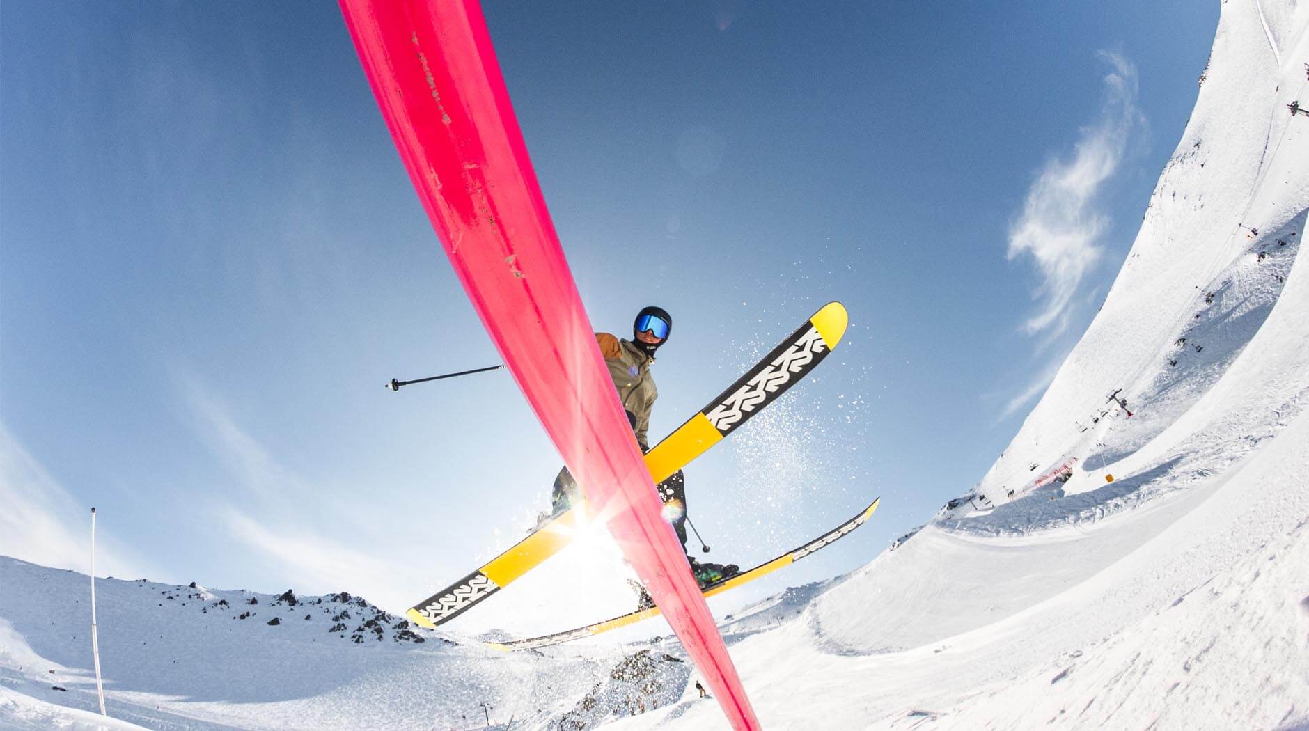 Skier jumping over a red rail on a snowy mountain under a clear blue sky.