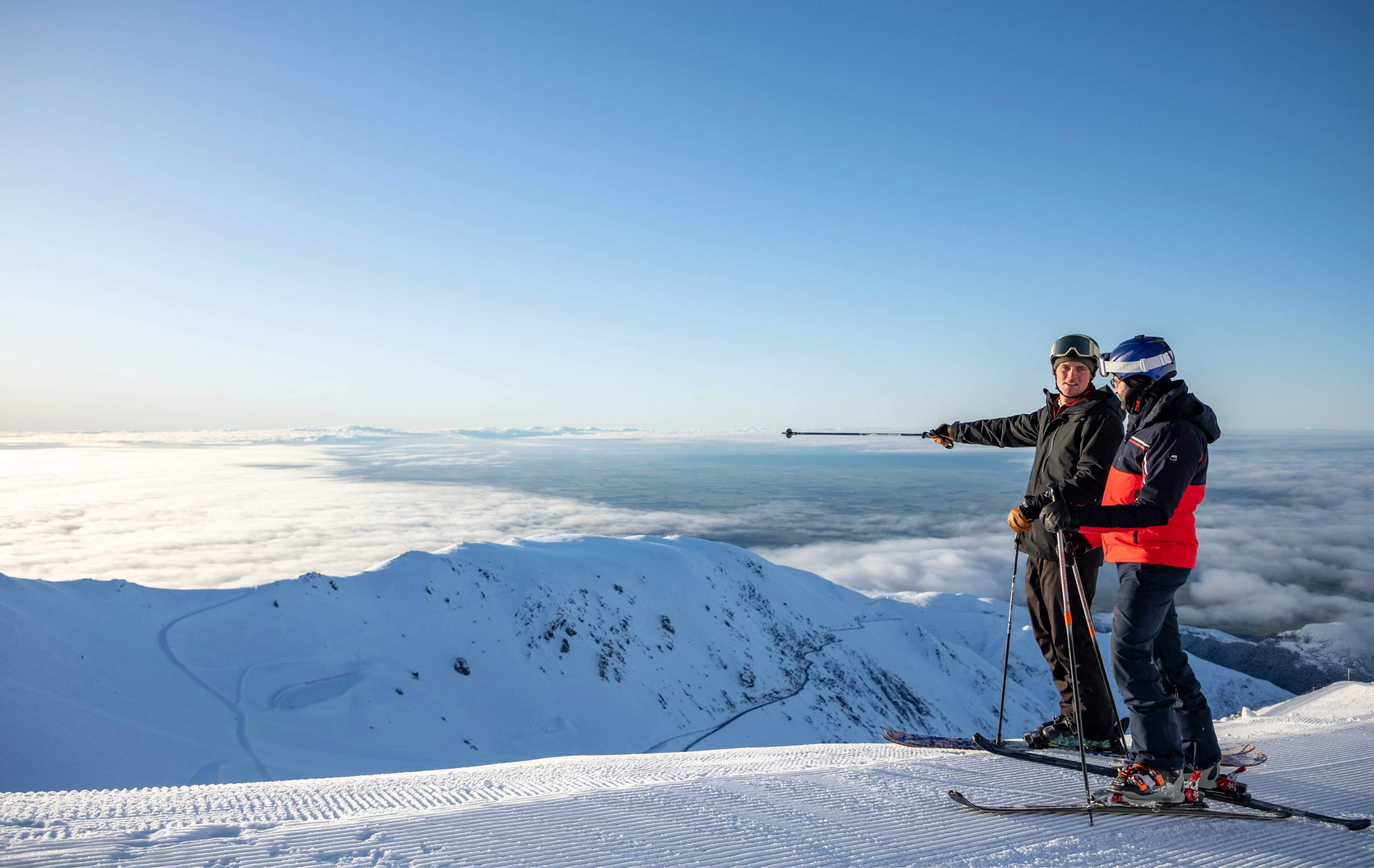 Two skiers standing on a snowy mountain peak, one pointing with a ski pole towards the distance above a sea of clouds under a clear blue sky.