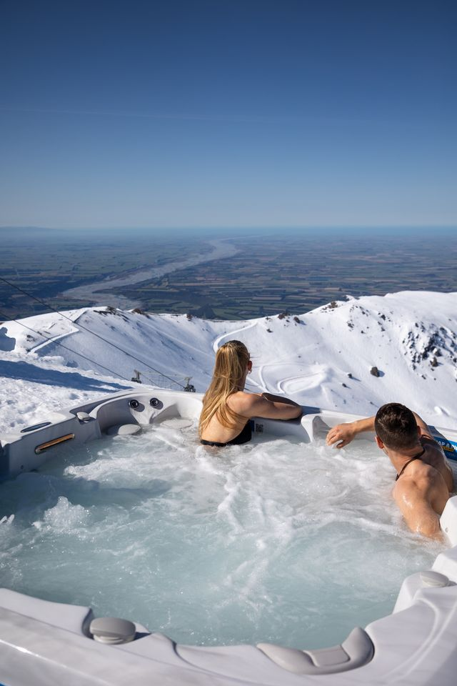 A man and woman relaxing in a hot tub on a snowy mountain with a clear view of a river and fields below.