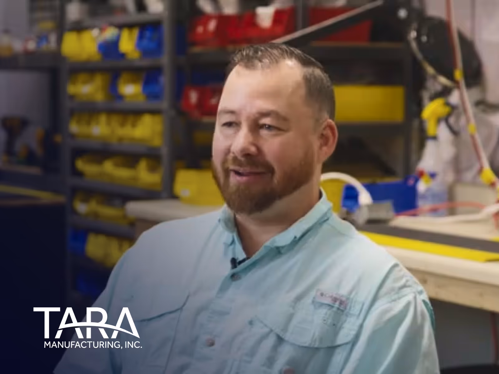 Man with a beard wearing a light blue button-up shirt sitting in a workshop with shelves of yellow and red bins behind him.