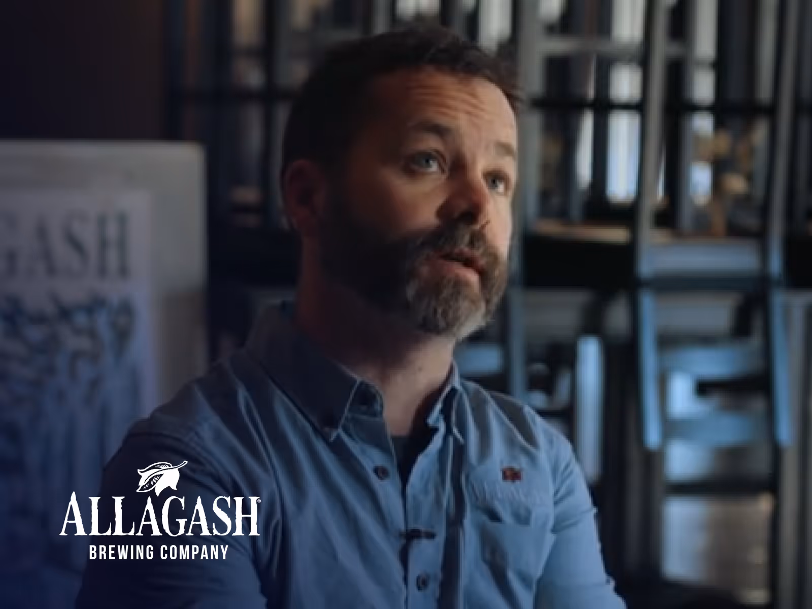 Man with beard wearing a blue shirt speaking indoors with stacked chairs in the background and Allagash Brewing Company logo.