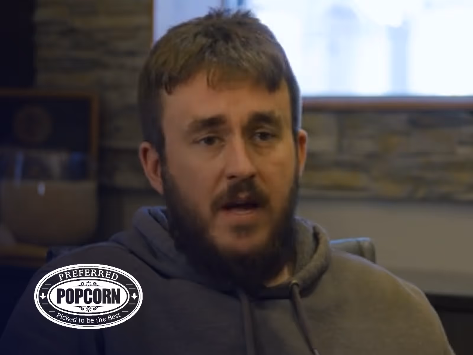 Bearded man in a hoodie speaking indoors with a 'Preferred Popcorn Picked to be the Best' logo in the bottom left corner.