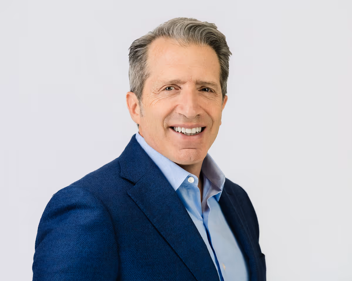 Mature man with gray hair smiling, wearing a blue suit jacket and light blue shirt, against a plain light background.