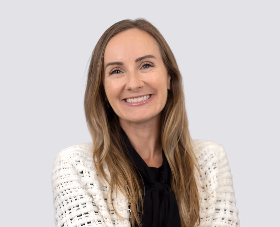 Smiling woman with long blonde hair wearing a white textured blazer and a black blouse against a plain light grey background.