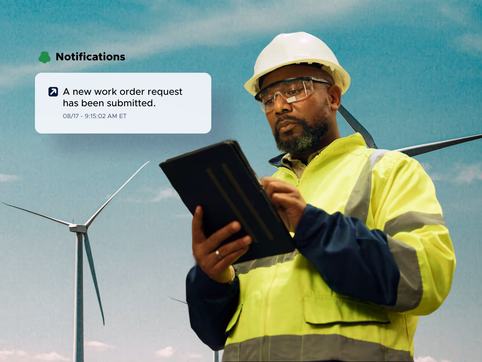 Engineer in safety gear using a tablet in front of wind turbines with notification about a new work order request.