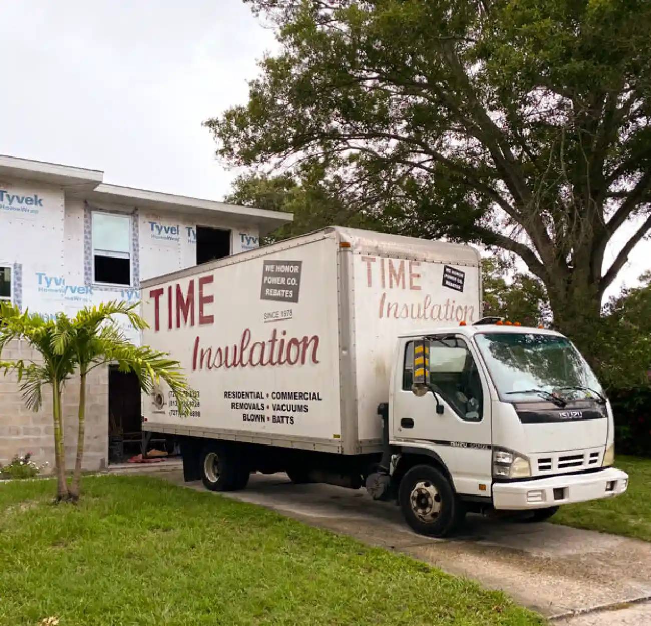 White Time Insulation truck parked on driveway next to a house under construction with Tyvek wrap and green lawn with a small palm tree.