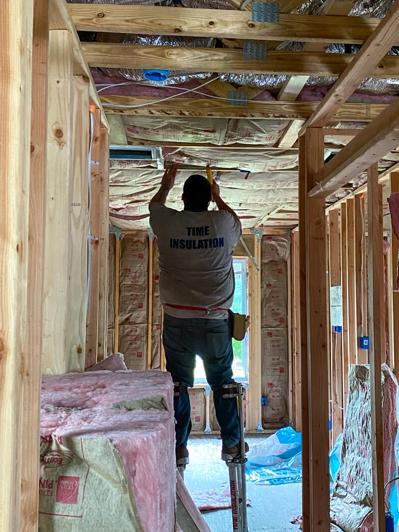 Worker on stilts installing insulation in the ceiling of a wooden framed house under construction.