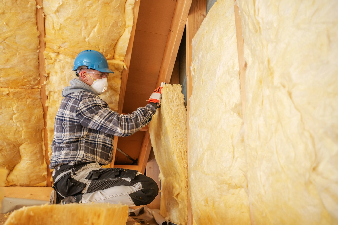 Worker wearing a blue hard hat and mask installing yellow insulation panels in an attic.