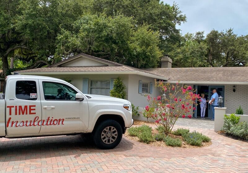 White pickup truck with 'TIME Insulation' logo parked in driveway of a one-story house where two people stand at an open front door.