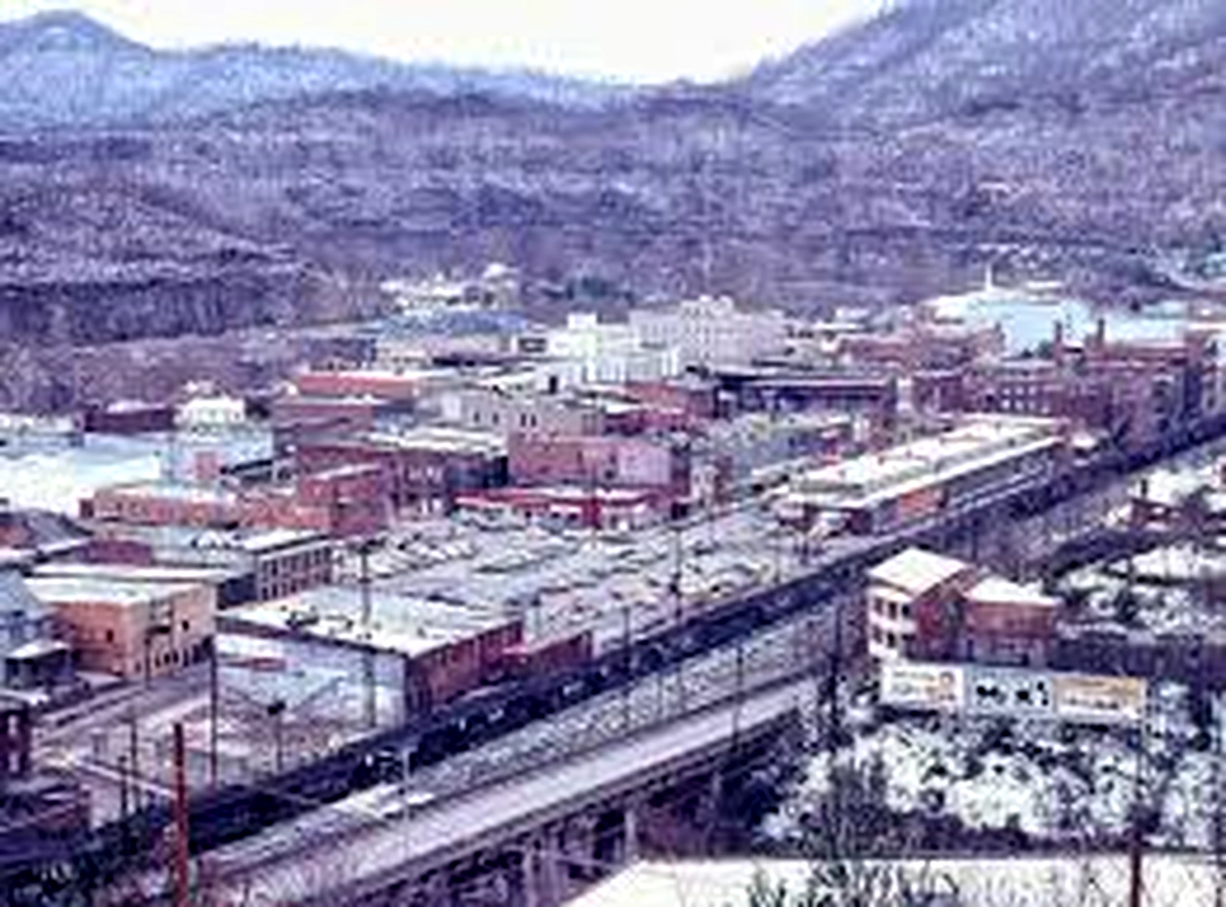 Panoramic view of an industrial town in a valley with buildings, a railway track, and surrounding hills.
