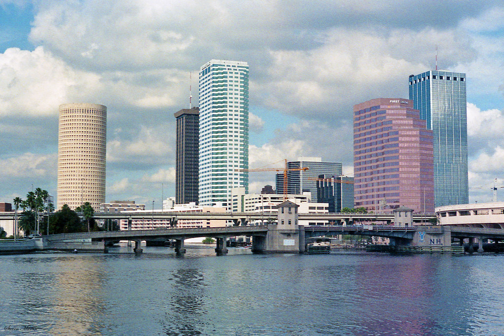 Daytime city skyline of Tampa, Florida, featuring several tall buildings behind a bridge over the water under a partly cloudy sky.