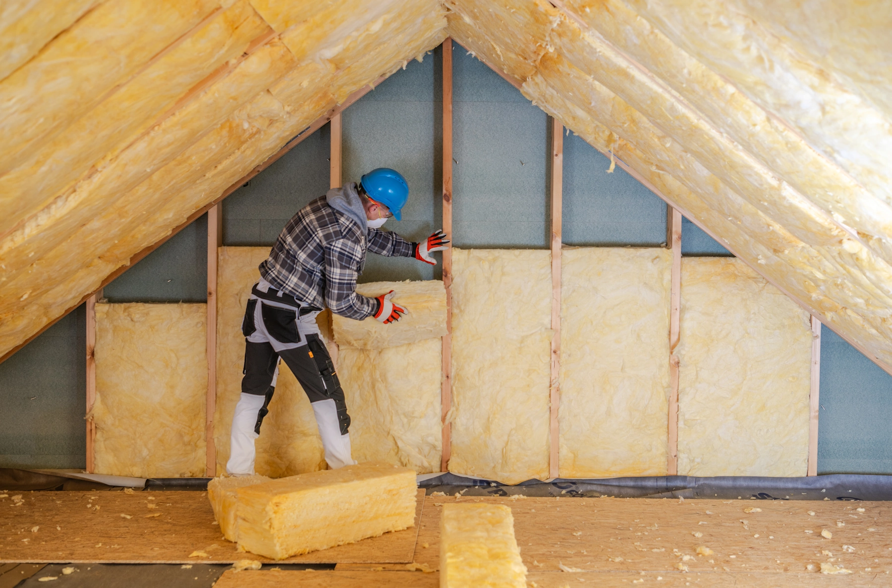 Worker wearing protective gear installing yellow batt insulation in an attic with wooden framework.