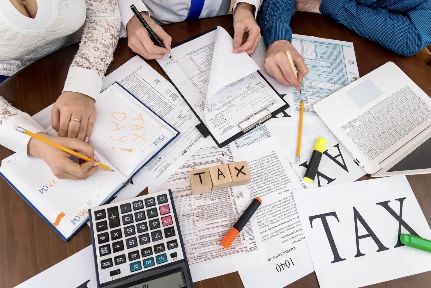 Group of people working together with calculators, tax forms, highlighters, and a notebook labeled "Tax Day" on a table.