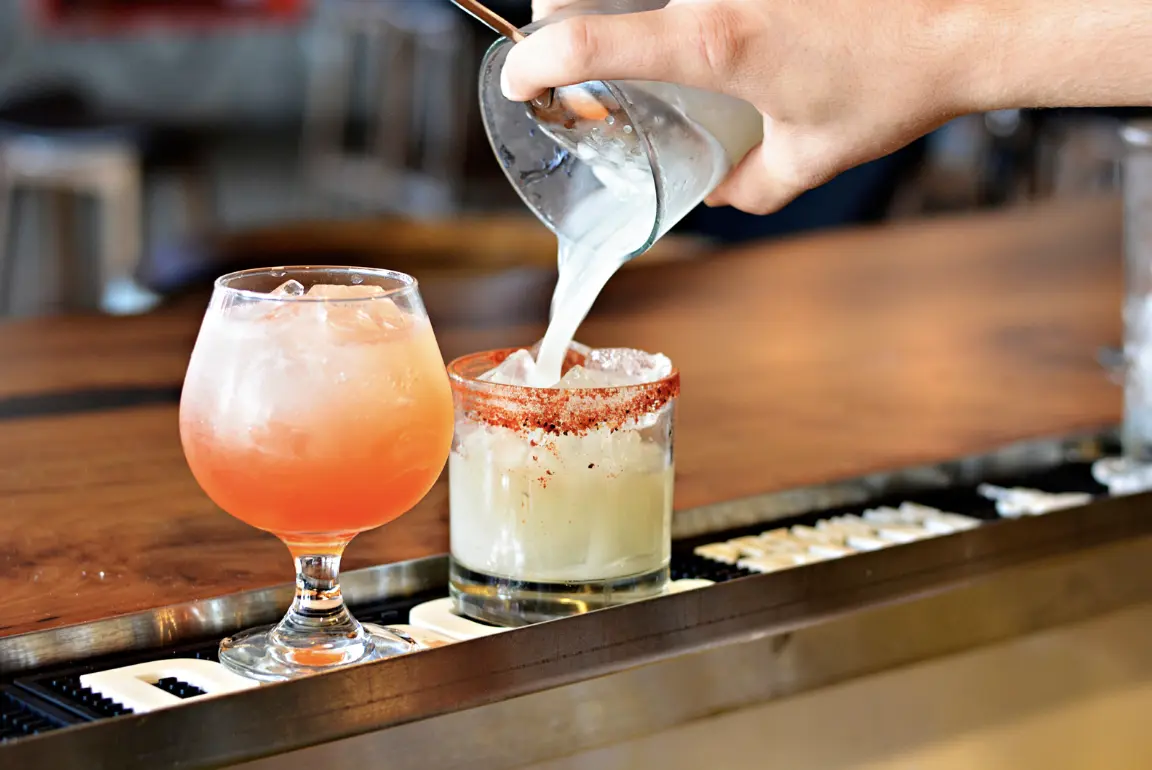 A close-up shot of a bartender pouring a creamy white liquid into a margarita glass, next to another vibrant cocktail with a salted rim.