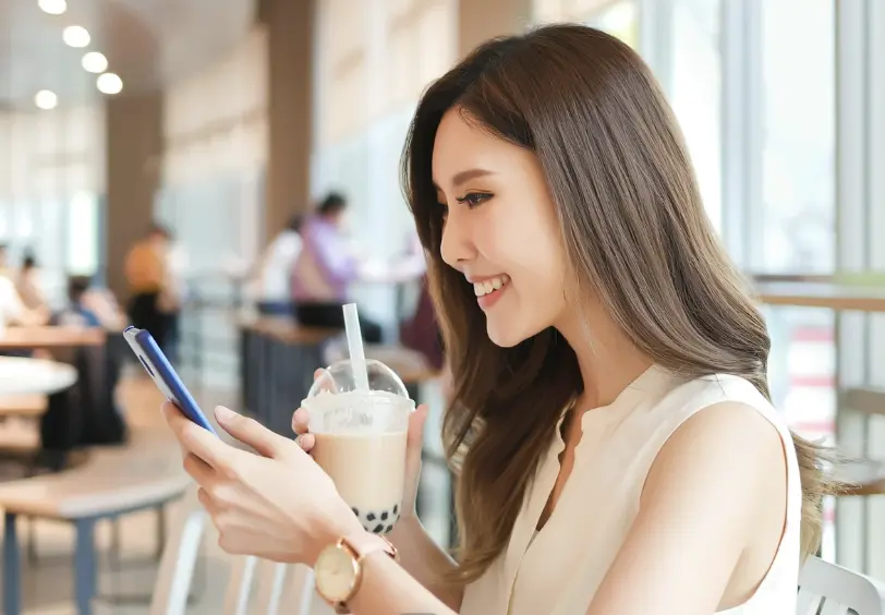 A smiling woman enjoying her bubble tea while checking her phone in a modern bubble tea shop, showcasing a seamless customer experience.