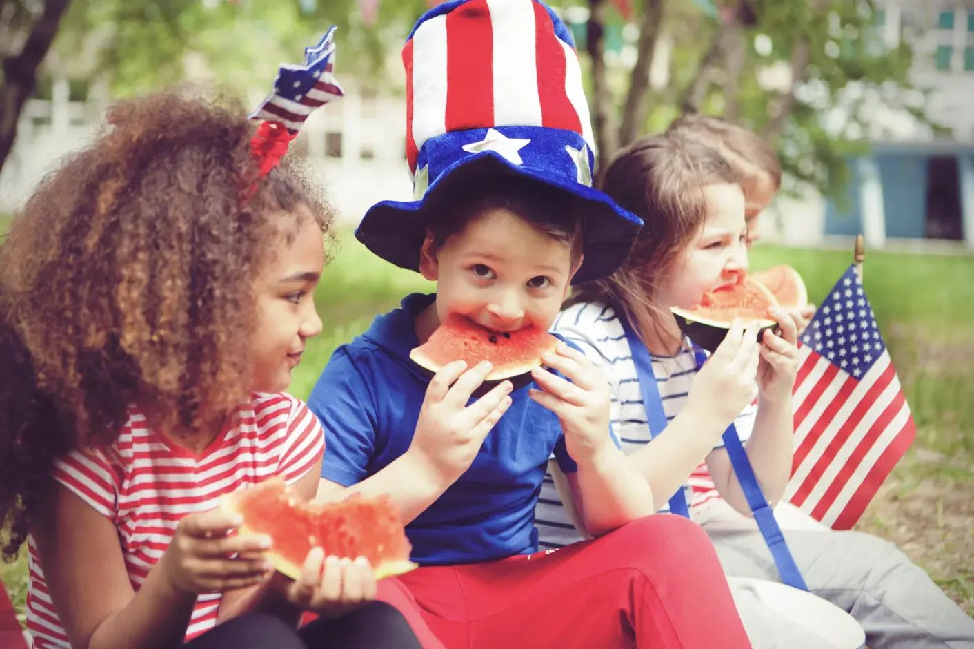 A group of young children in patriotic clothing eating watermelon during a 4th of July picnic, with flags and festive accessories.