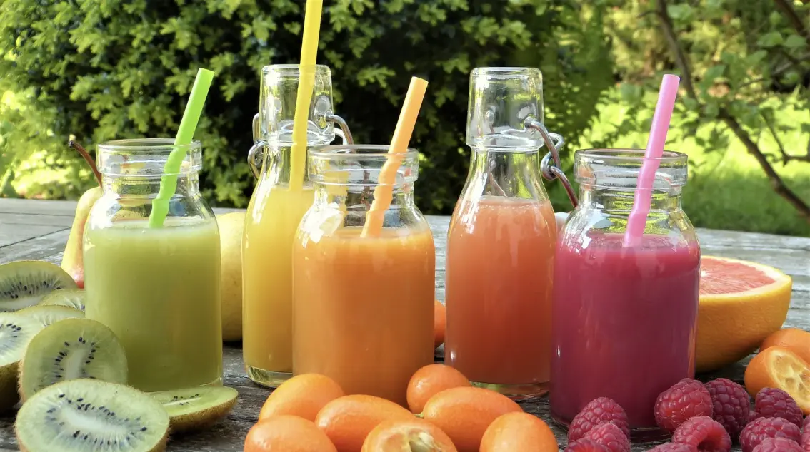 A variety of colorful fresh fruit juices in glass bottles with straws, surrounded by kiwis, kumquats, raspberries, and grapefruits on a wooden table.