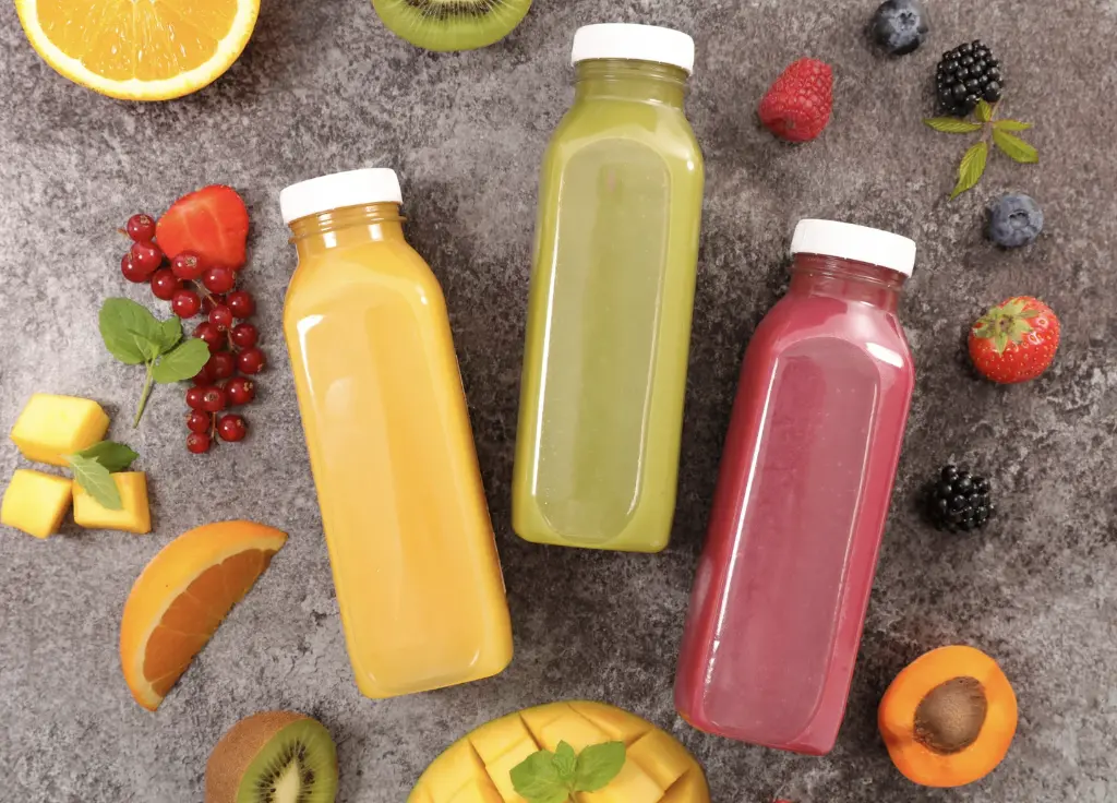 Three colorful bottled fruit juices in yellow, green, and pink, surrounded by fresh fruits like oranges, mangoes, berries, and kiwis on a textured gray surface.