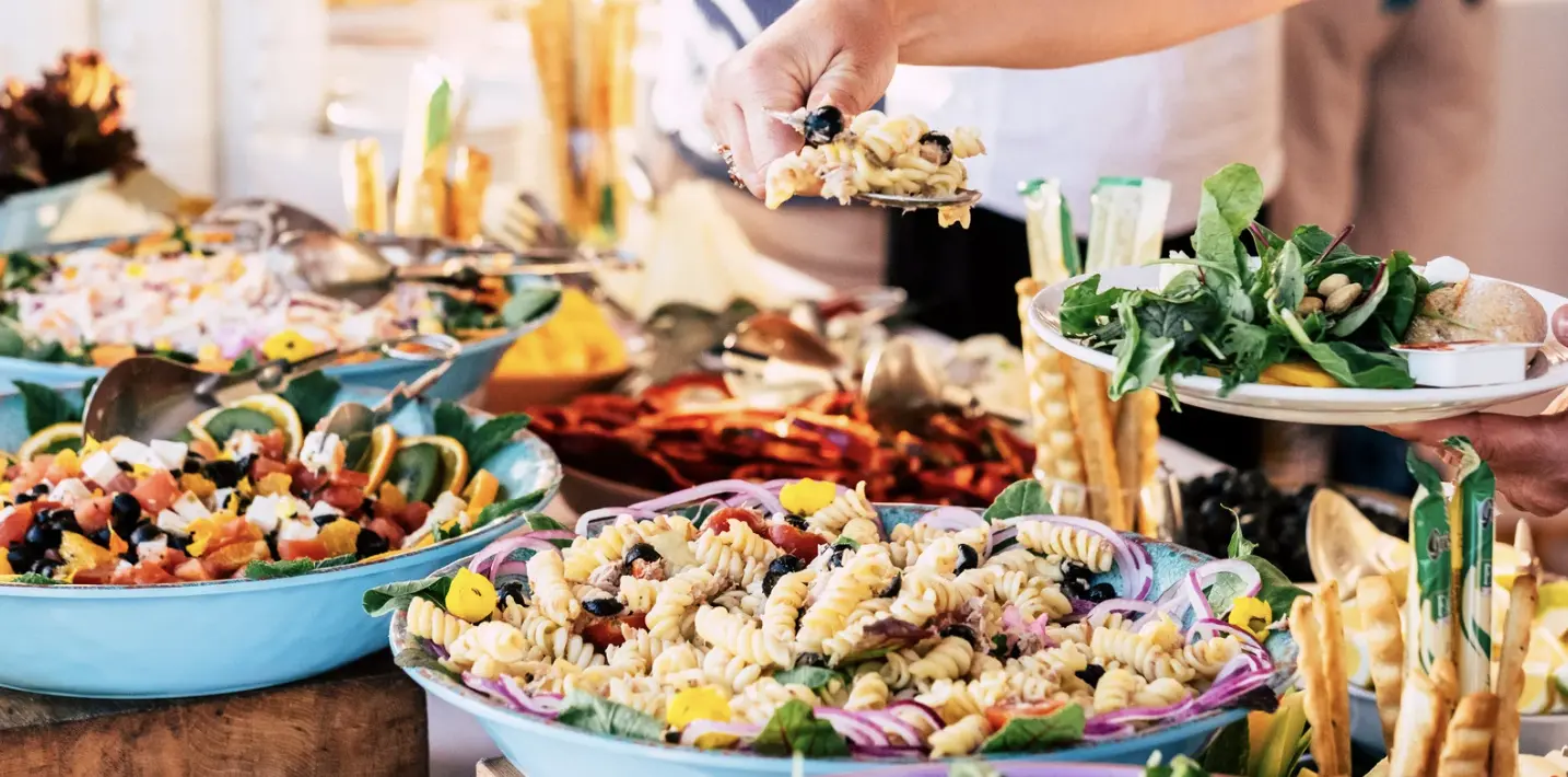 A hand serving pasta salad from a vibrant buffet display featuring leafy greens, citrus fruits, and colorful ingredients in large blue bowls.