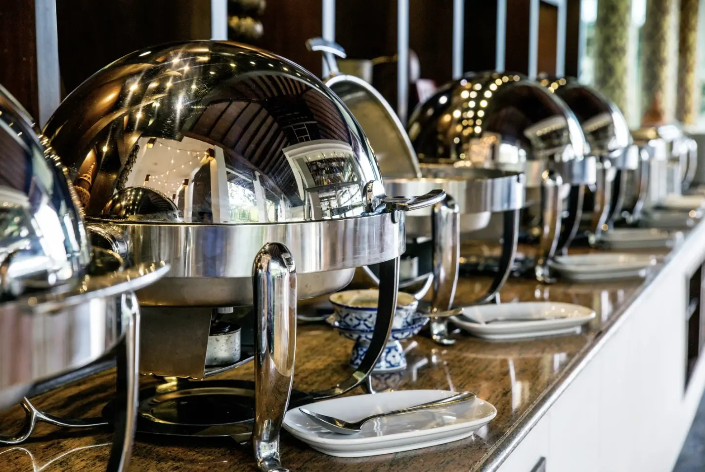 Row of shiny stainless steel chafing dishes with lids closed, lined up on a polished countertop in a high-end buffet setting.