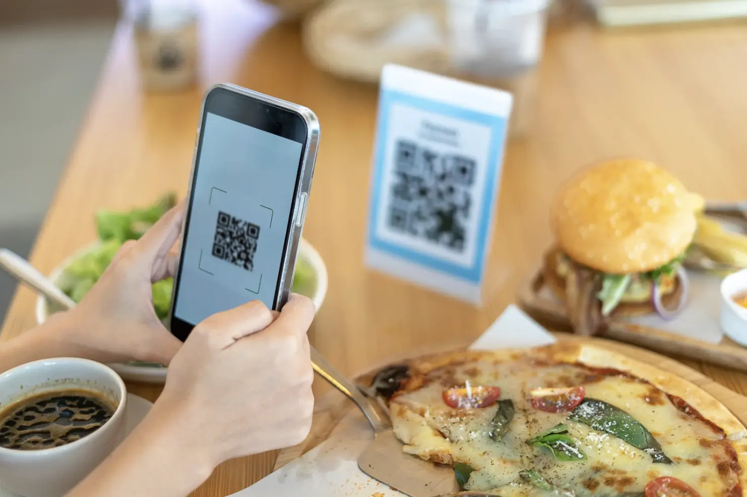 A customer scanning a QR code menu on a table next to pizza, salad, and coffee at a casual dining spot.