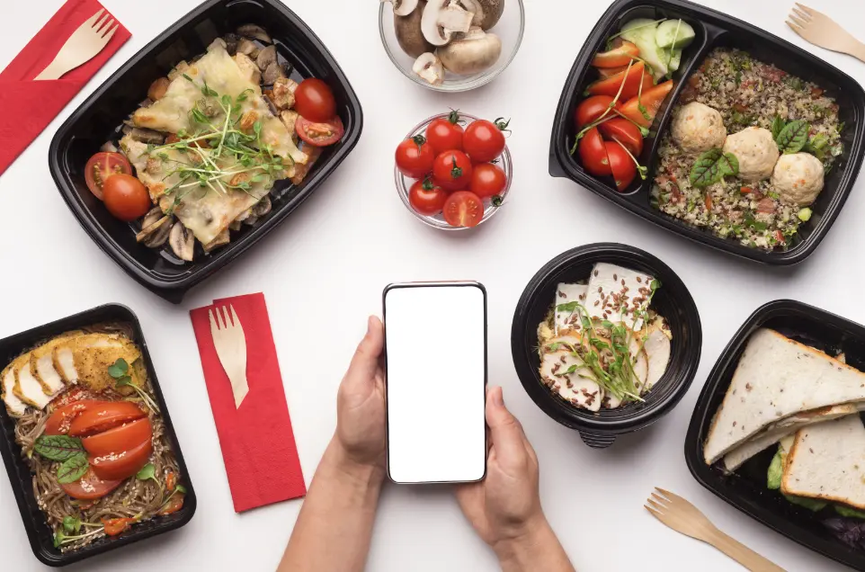 A person using a smartphone to order food, surrounded by a variety of takeout meals, highlighting AI-driven food service.