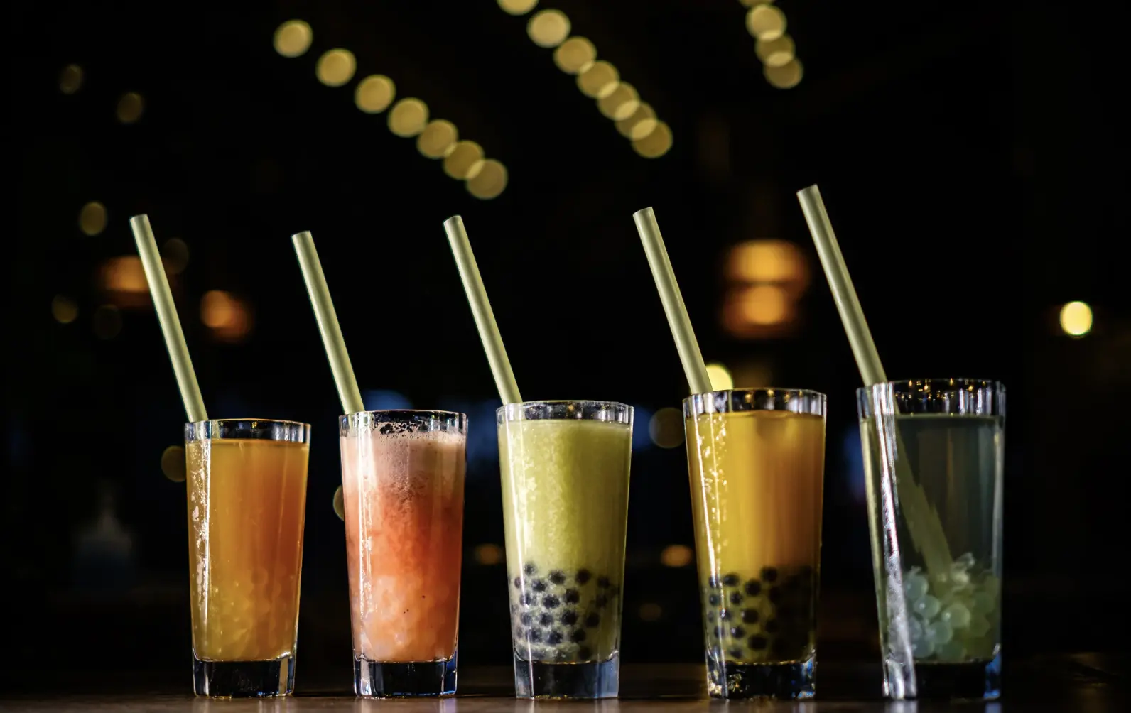 Five vibrant bubble tea drinks with different colors and toppings lined up on a table in a warmly lit cafe.