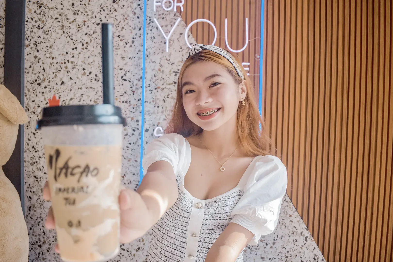 A cheerful young woman holding out her bubble tea drink, smiling towards the camera in a stylish bubble tea shop.