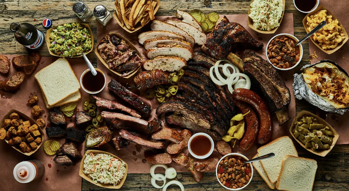 A large BBQ feast featuring smoked ribs, sausage, chicken, brisket, and sides like coleslaw, baked beans, mac and cheese, and pickles.