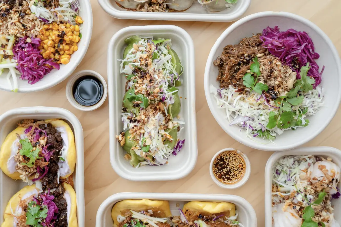 Assorted takeout dishes including bao buns, rice bowls, and vibrant vegetables in eco-friendly containers.