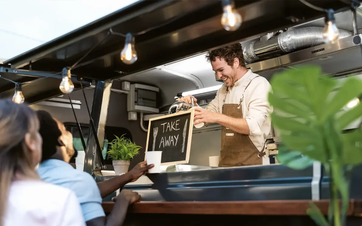Smiling food truck vendor handing food to customers under string lights at a takeout window.
