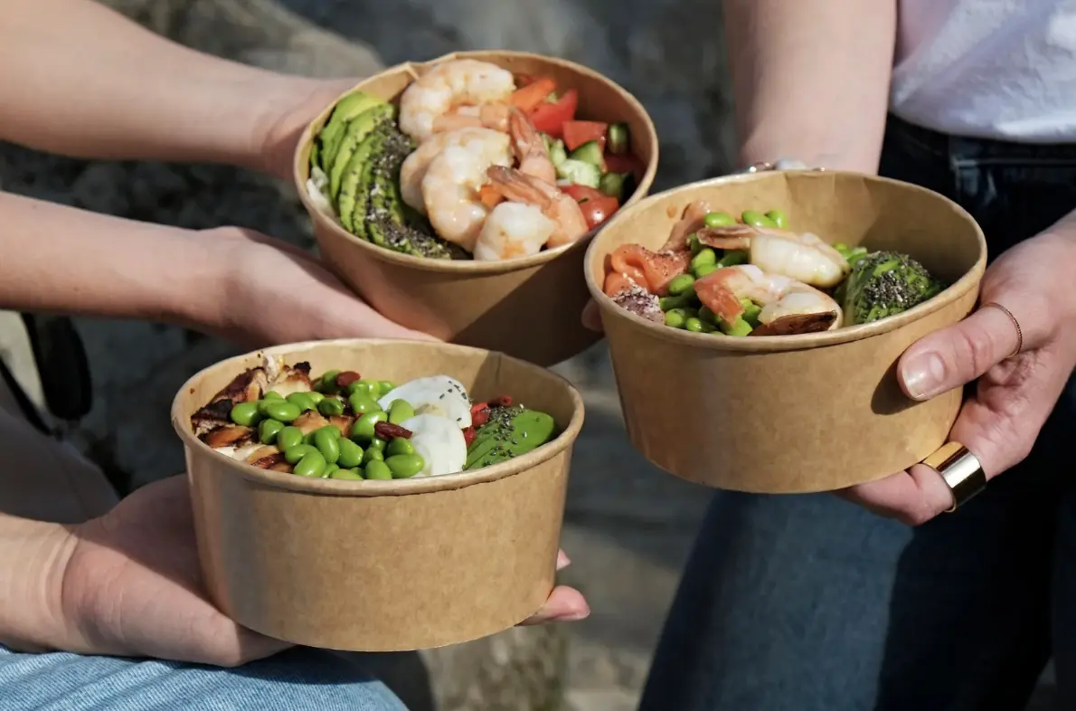 Three hands holding paper bowls filled with shrimp, avocado, edamame, and colorful toppings outdoors.