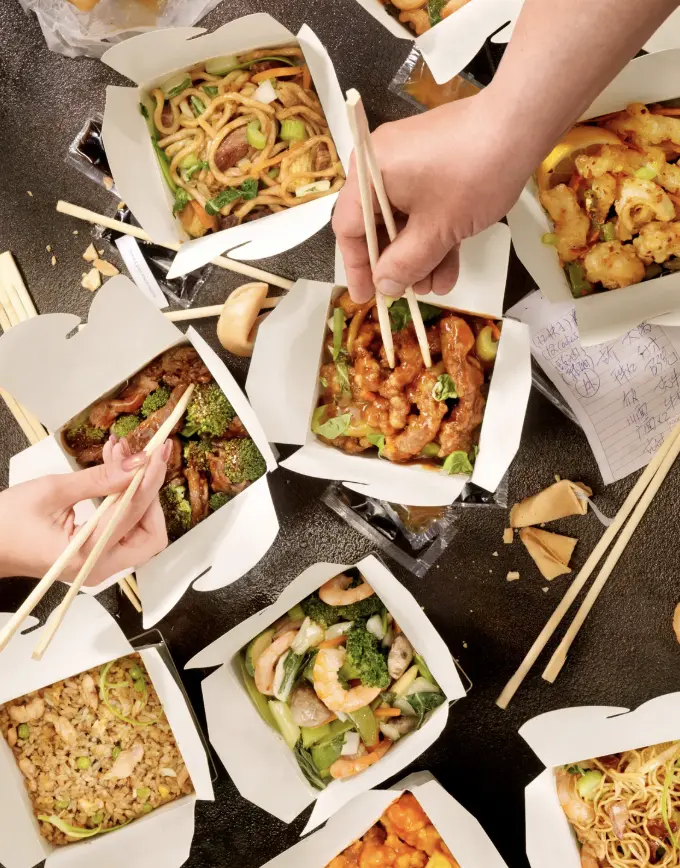 Hands using chopsticks to enjoy various Chinese takeout dishes in iconic white paper boxes.