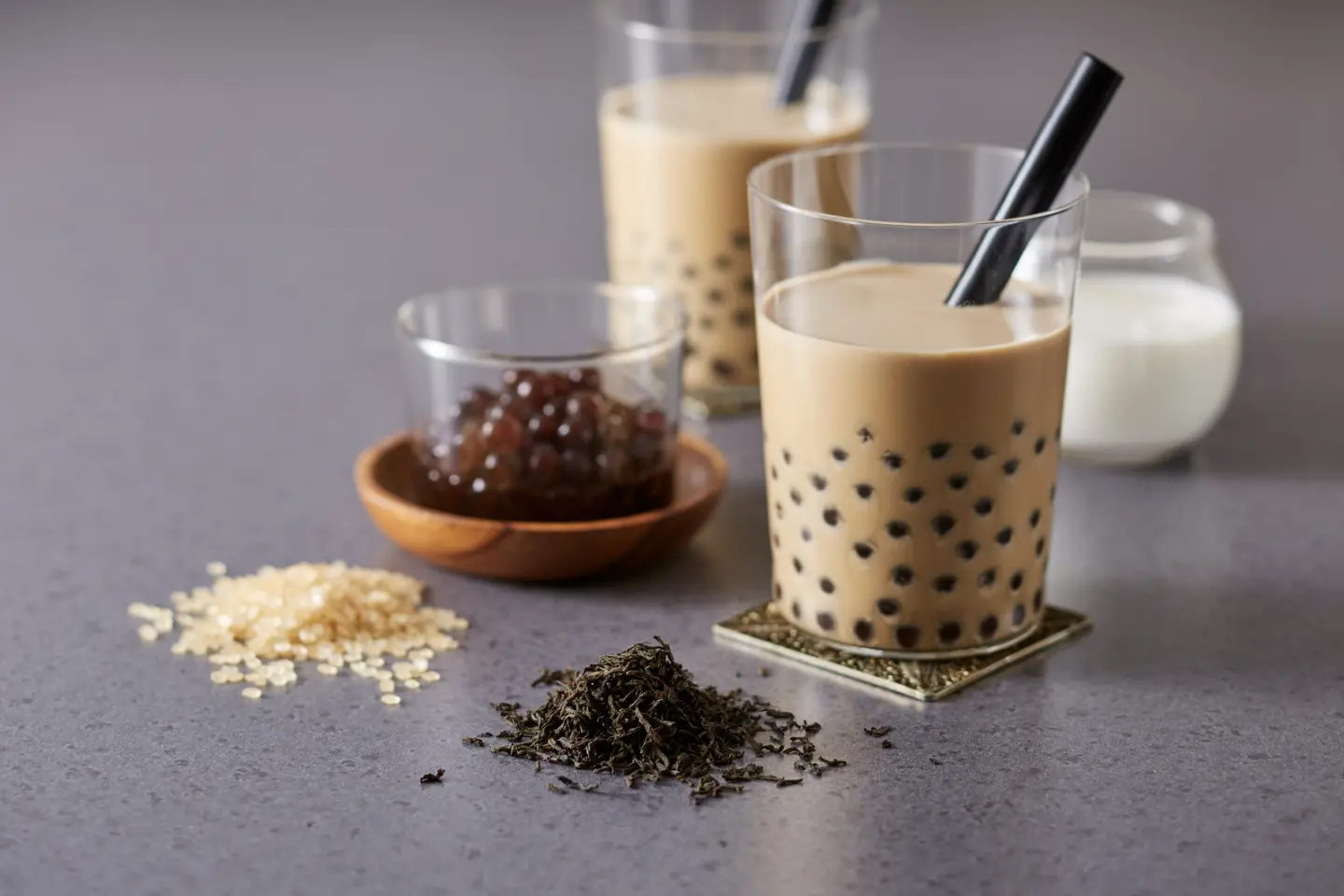 Close-up of bubble tea ingredients including tapioca pearls, tea leaves, sugar, and fresh milk with two glasses of bubble tea.