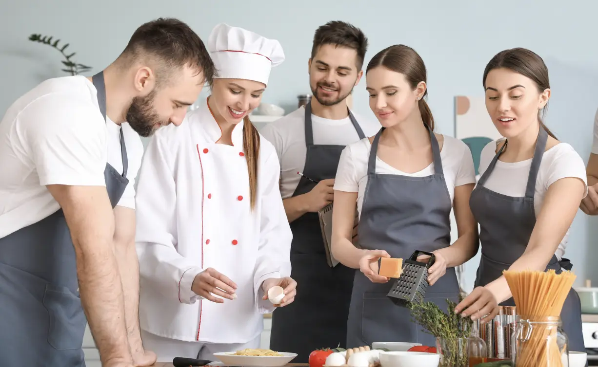 A pastry chef in a white uniform demonstrating techniques to a group of kitchen trainees in aprons.