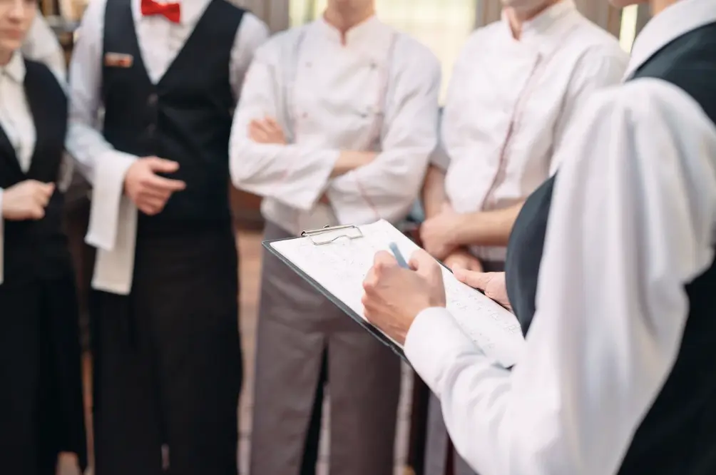 A restaurant manager holding a clipboard while discussing operations and planning with the kitchen and waitstaff team.