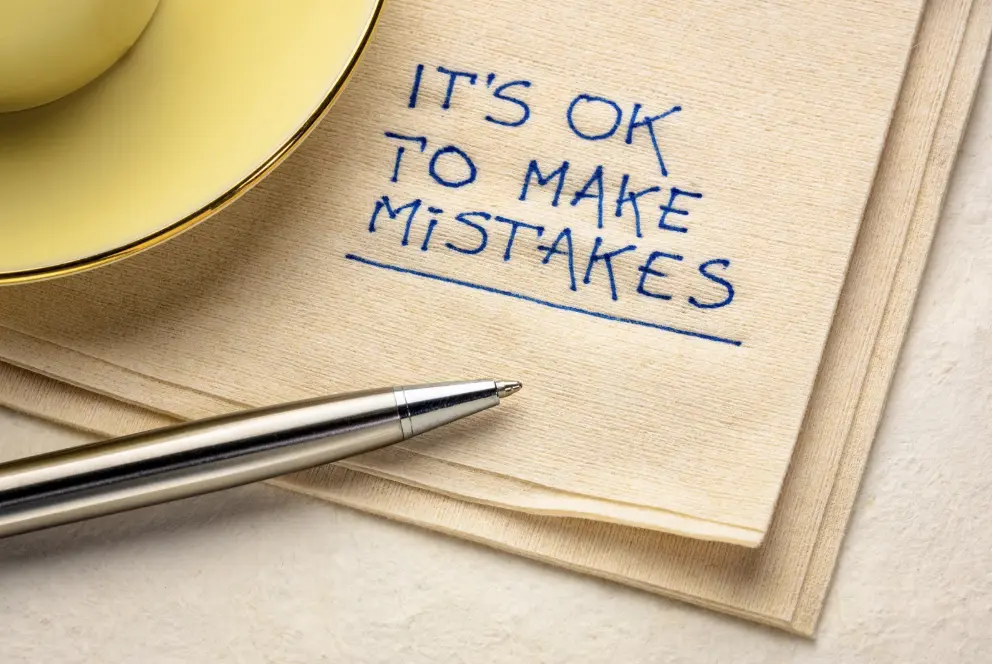 A napkin with handwritten text "It's OK to make mistakes," placed beside a pen and a yellow cup, suggesting acceptance of errors.