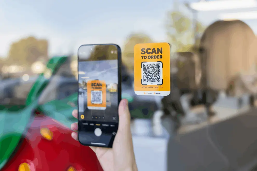 Person scanning a yellow "Scan to Order" QR code sign on a window using a smartphone camera.