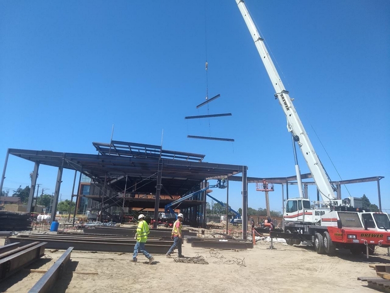 Construction workers guide structural steel beams being lifted by a crane during erection of a steel-framed building