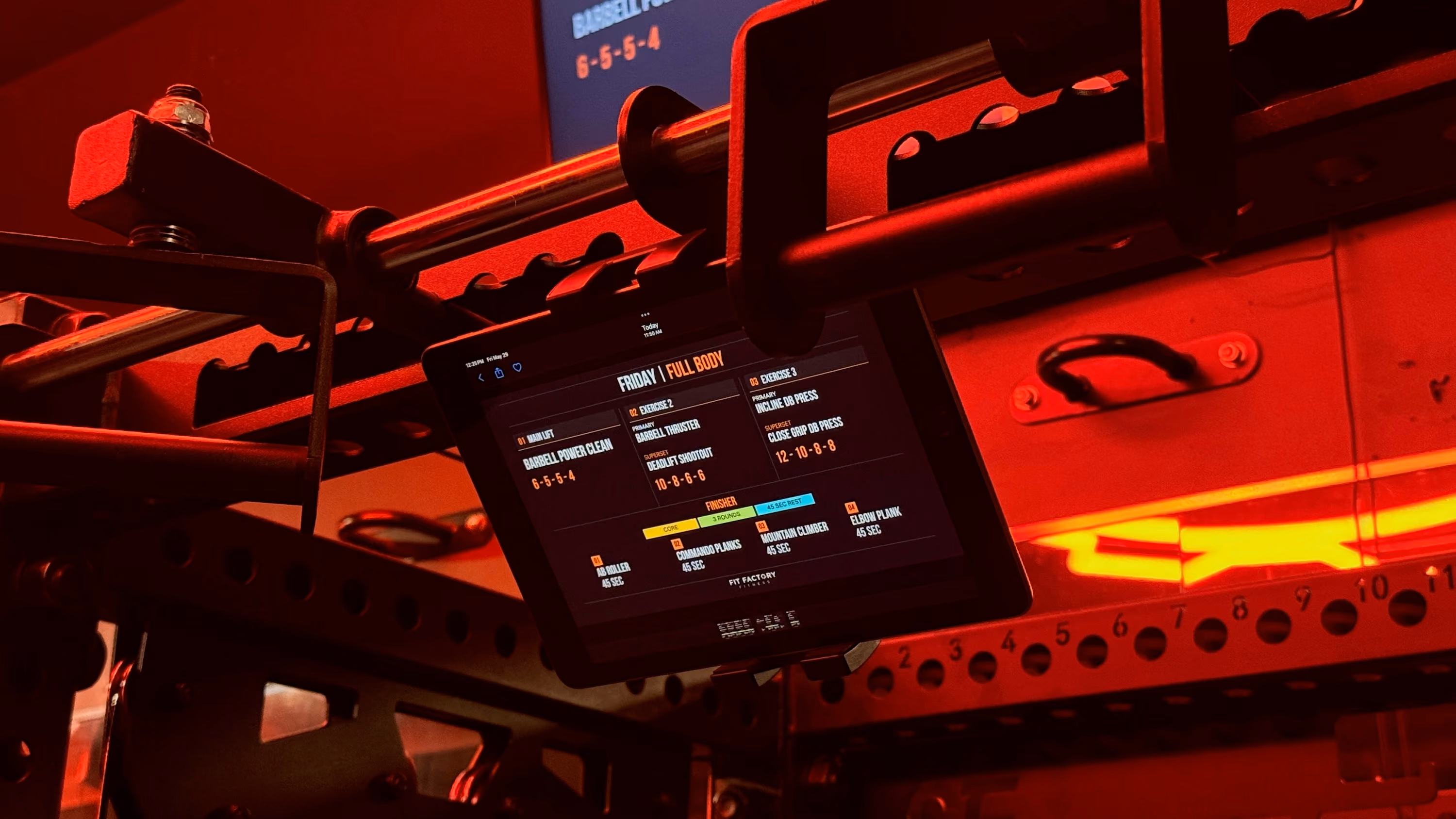 Workout programming screen mounted on a squat rig on the Fit Factory strength floor, lit red