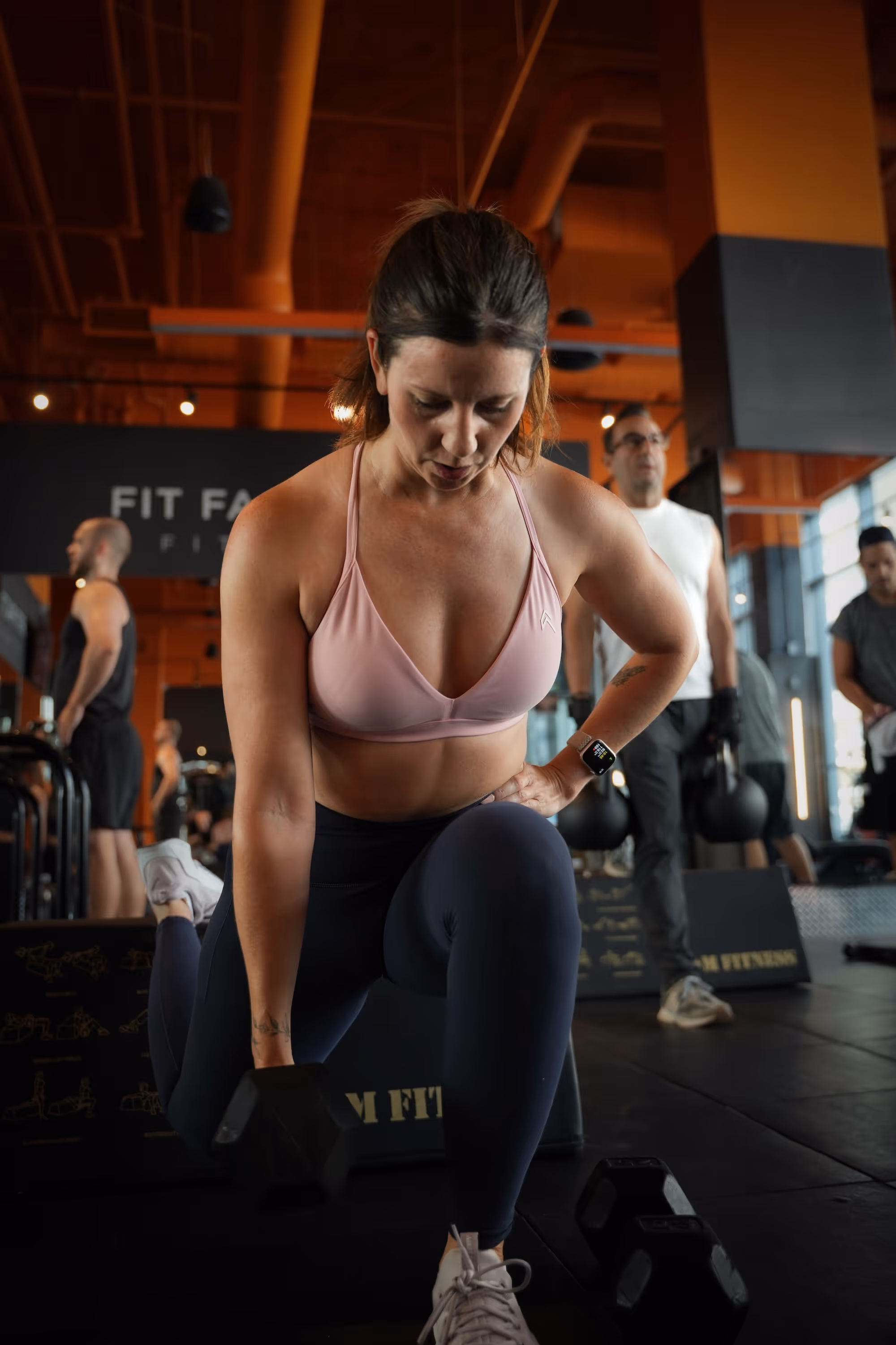 Member in a split squat holding a kettlebell at her chest, with other members training in the background