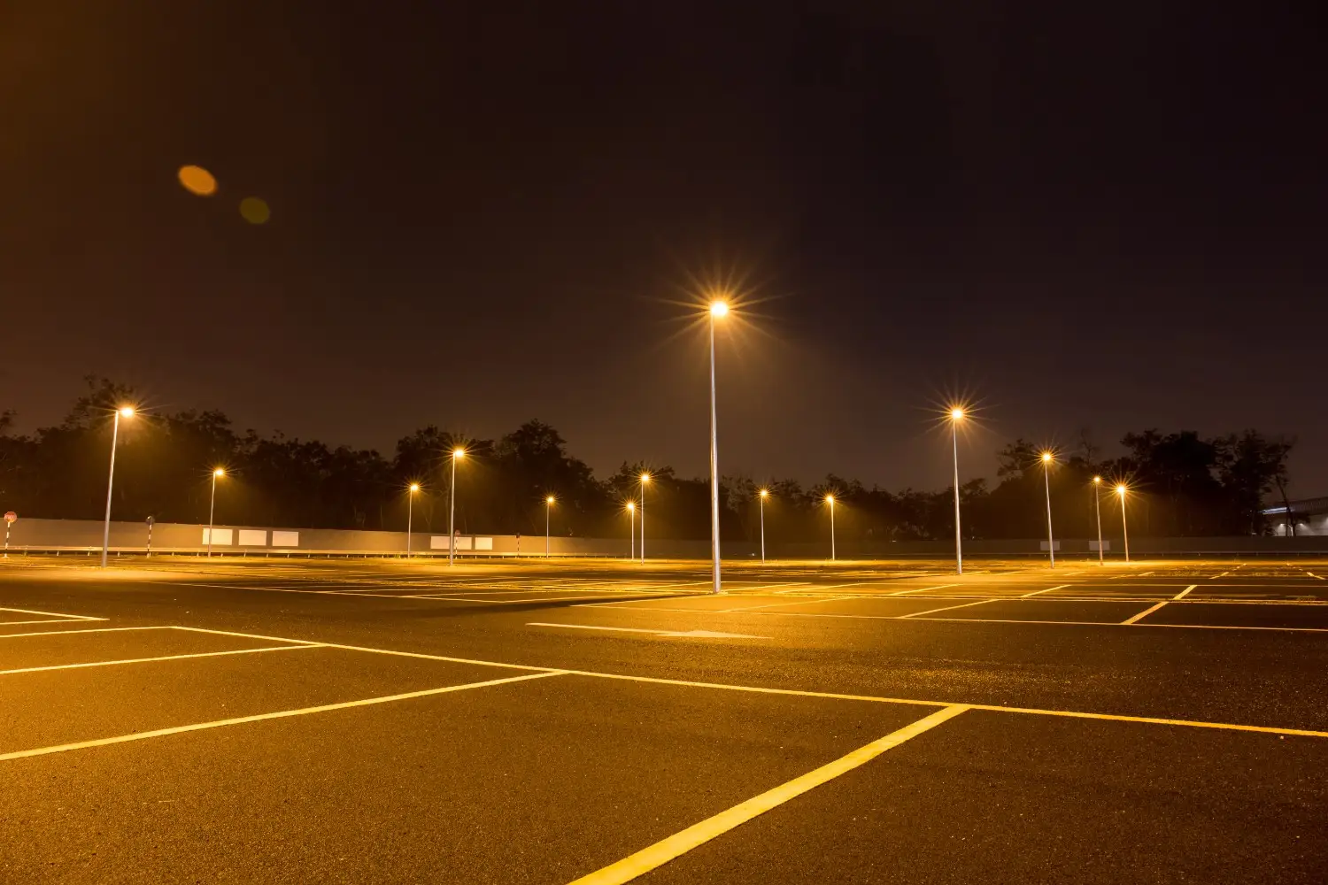 An empty parking lot at night illuminated by several tall LED pole lights casting a warm orange glow across the pavement.