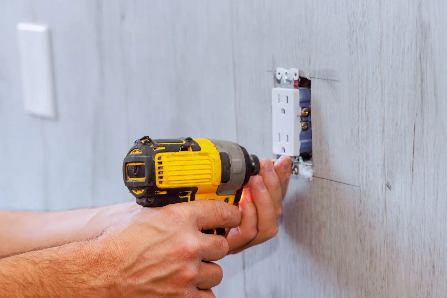 Close-up of hands using a yellow power drill to install a white electrical outlet into a light wood-paneled wall.