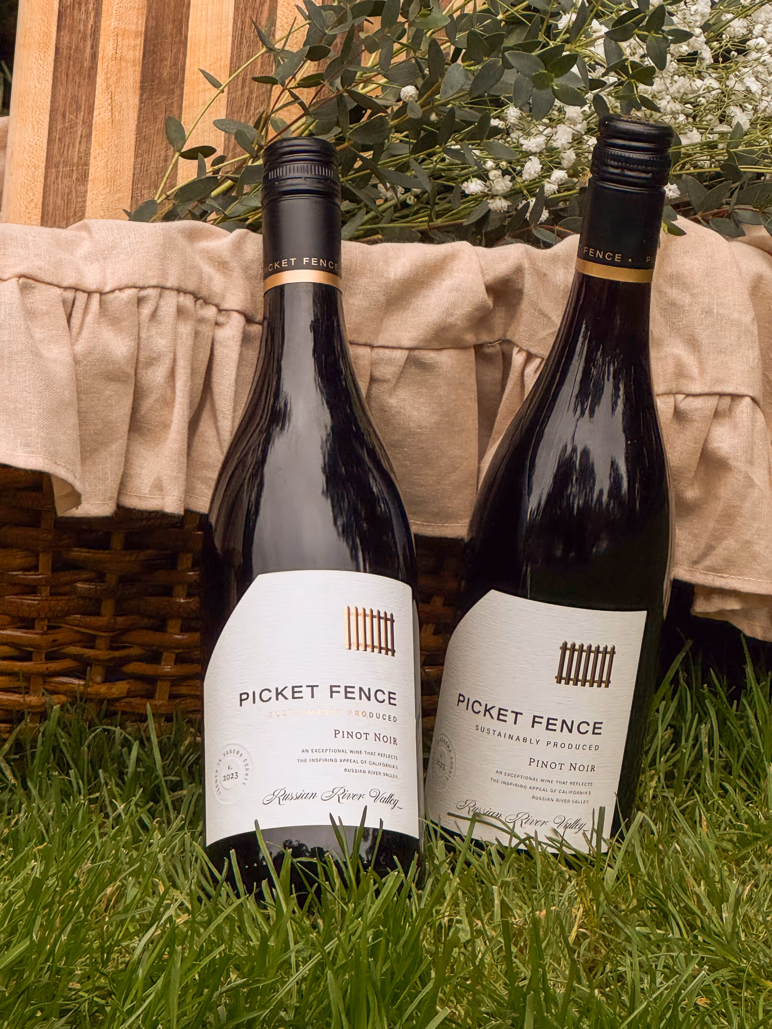 Two bottles of Picket Fence Pinot Noir wine placed on grass in front of a wicker basket with beige cloth and floral greenery.
