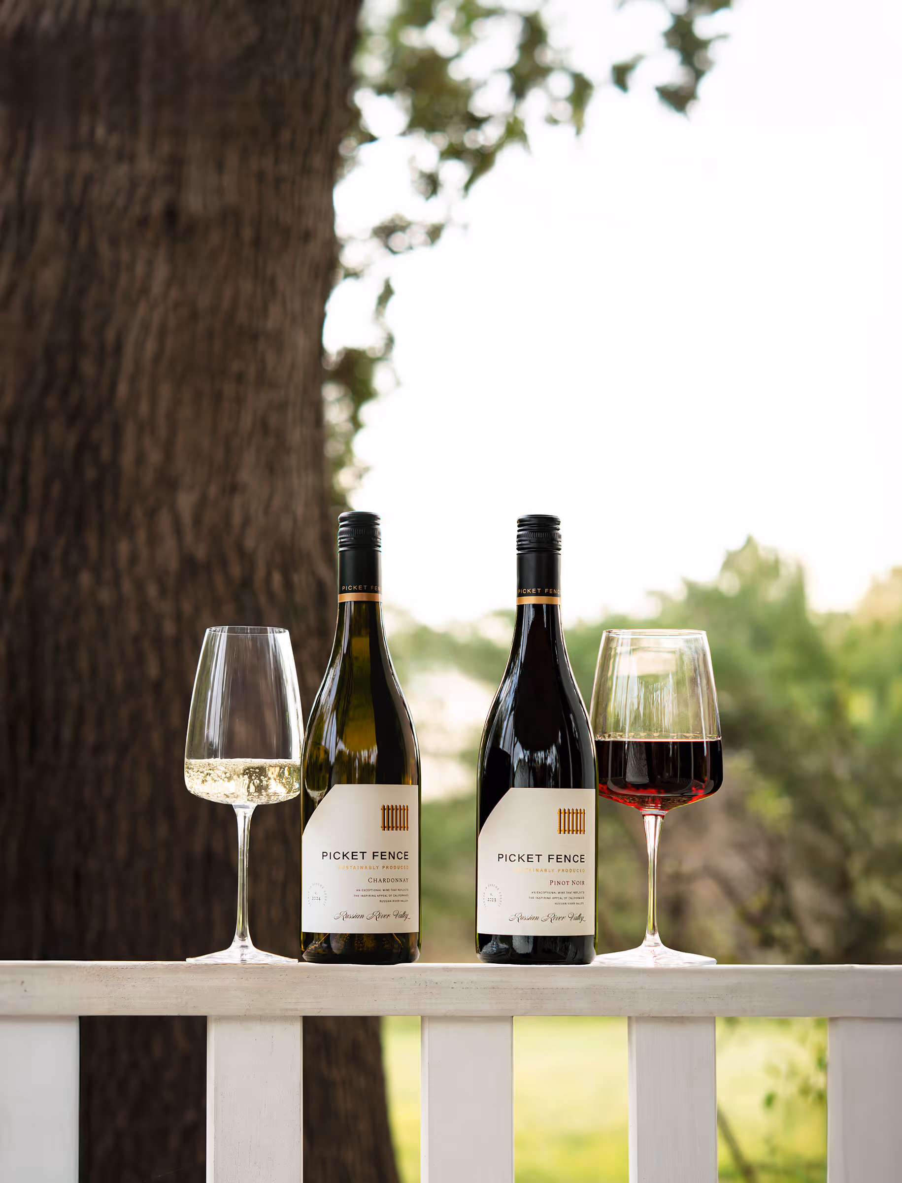 Two bottles of Picket Fence wine, Chardonnay and Pinot Noir, with matching filled wine glasses on a white wooden railing outdoors.