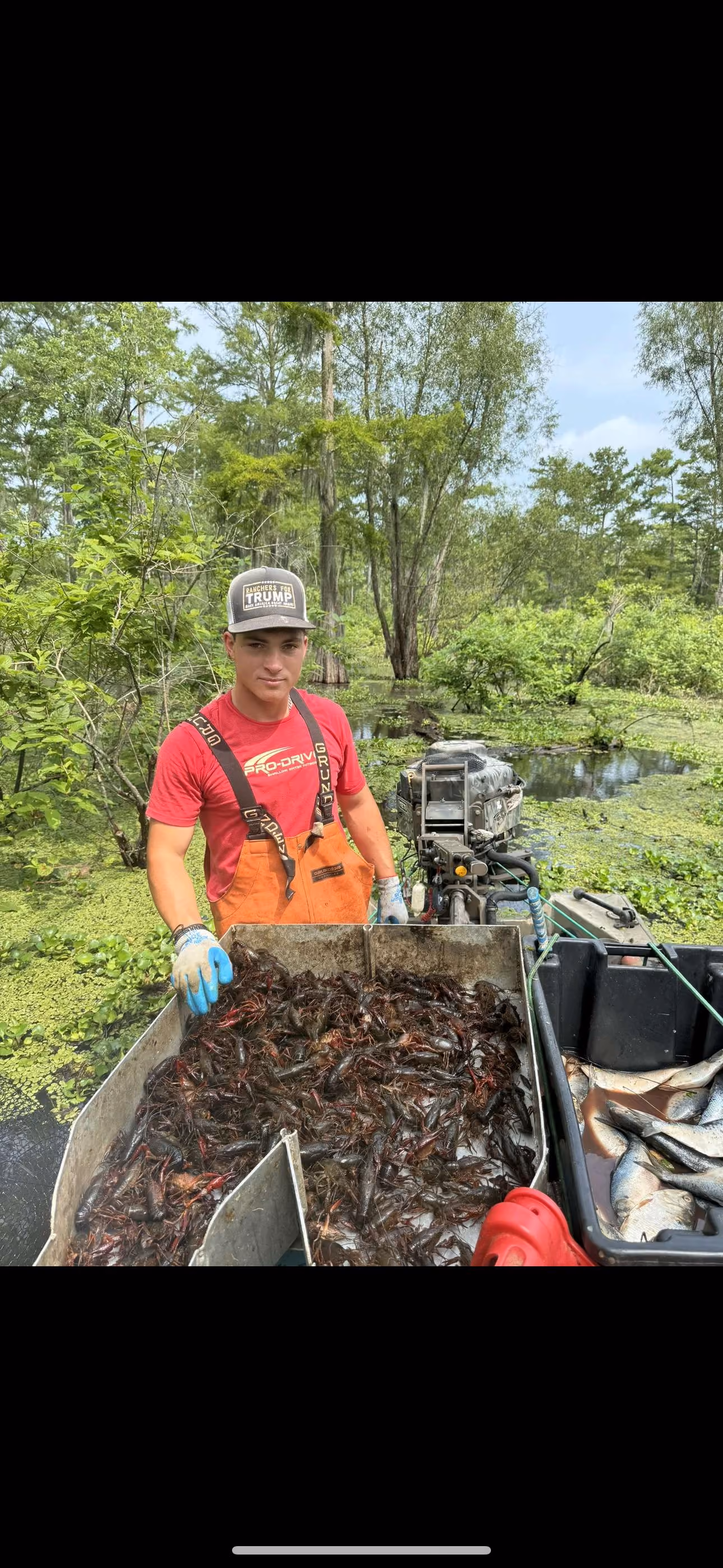 Man in fishing gear holding a container full of crawfish on a boat in a swampy, green water area with fish in a black container beside him.